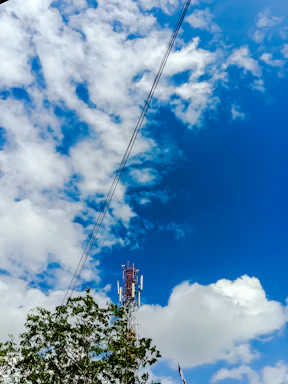 A telecommunications tower stands tall against a vibrant blue sky scattered with fluffy white clouds. Green foliage and overhead power lines frame the scene, adding a contrasting natural element to the industrial structure of the tower.