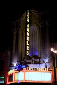A vintage-style theater facade is illuminated at night with bright neon lights displaying the name Los Angeles in bold letters. The structure features ornate architectural details, with intricate carvings and columns. The marquee at the bottom is lined with red and blue neon lights.