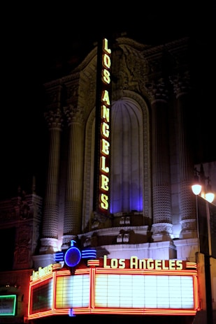 A vintage-style theater facade is illuminated at night with bright neon lights displaying the name Los Angeles in bold letters. The structure features ornate architectural details, with intricate carvings and columns. The marquee at the bottom is lined with red and blue neon lights.