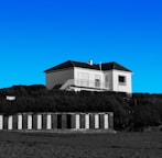 A white two-story house with a dark roof sits atop a grassy hill under a clear blue sky. Below the hill, there are a series of black and white striped beach cabanas. The scene appears calm and serene.