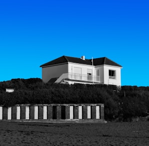 A white two-story house with a dark roof sits atop a grassy hill under a clear blue sky. Below the hill, there are a series of black and white striped beach cabanas. The scene appears calm and serene.