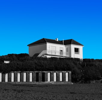 A white two-story house with a dark roof sits atop a grassy hill under a clear blue sky. Below the hill, there are a series of black and white striped beach cabanas. The scene appears calm and serene.