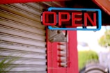 A brightly lit neon sign displaying the word 'OPEN' hangs against a partially visible metallic shutter. The sign is outlined with blue and features red lettering. There is a red structural beam in the background along with some blurred green foliage.
