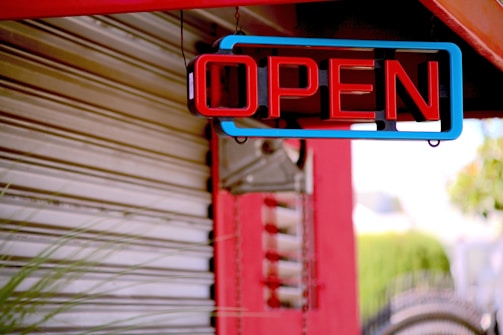 A brightly lit neon sign displaying the word 'OPEN' hangs against a partially visible metallic shutter. The sign is outlined with blue and features red lettering. There is a red structural beam in the background along with some blurred green foliage.