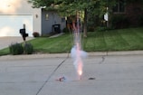 A firework is set up on a residential street, emitting sparks and smoke into the air. The background includes a well-maintained house with a garage, a driveway, and a green lawn. There are flowers and neatly trimmed shrubs lining the house, and a mailbox is positioned near the curb. Trees partially shade the area, and sunlight casts shadows across the scene.