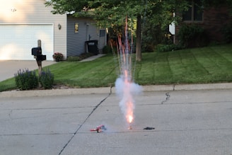 A firework is set up on a residential street, emitting sparks and smoke into the air. The background includes a well-maintained house with a garage, a driveway, and a green lawn. There are flowers and neatly trimmed shrubs lining the house, and a mailbox is positioned near the curb. Trees partially shade the area, and sunlight casts shadows across the scene.