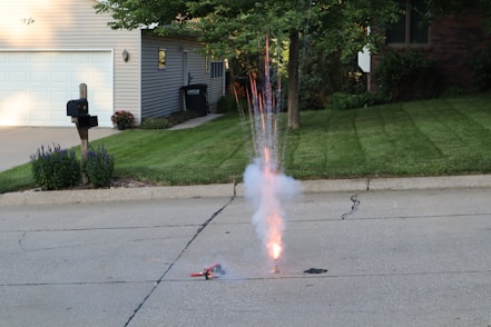 A firework is set up on a residential street, emitting sparks and smoke into the air. The background includes a well-maintained house with a garage, a driveway, and a green lawn. There are flowers and neatly trimmed shrubs lining the house, and a mailbox is positioned near the curb. Trees partially shade the area, and sunlight casts shadows across the scene.