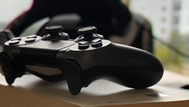 A black gaming controller is placed on top of a book in a softly lit environment. The controller's buttons and analog sticks are clearly visible, and the background is slightly blurred with hints of greenery.