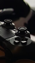 A close-up view of a black gaming controller, highlighting the joystick and button layout. The lighting is dim, creating a sleek and focused atmosphere.