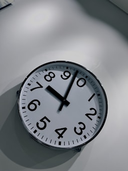 A wall clock with a white face and black numerals is mounted on a light-colored wall. The clock hands indicate the time is slightly past nine o&rsquo;clock. The setting appears minimalistic with an emphasis on simplicity.