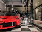 A sleek red sports car is prominently displayed inside a showroom with a modern and luxurious design. Next to it, a vintage motorcycle adds a classic touch to the space. The floor features a black and white checkered pattern, and large glass windows provide a view of the interior decor, which includes sophisticated lighting and wall art.