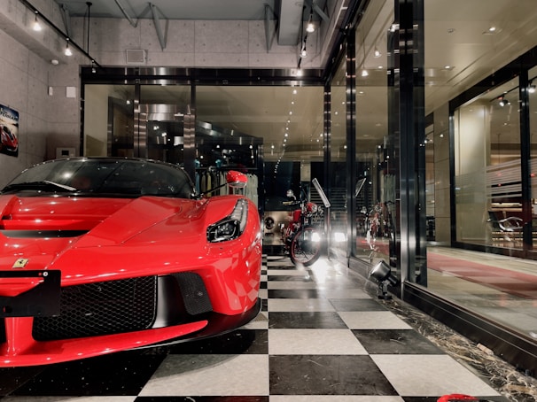 A sleek red sports car is prominently displayed inside a showroom with a modern and luxurious design. Next to it, a vintage motorcycle adds a classic touch to the space. The floor features a black and white checkered pattern, and large glass windows provide a view of the interior decor, which includes sophisticated lighting and wall art.