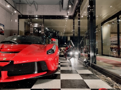 A sleek red sports car is prominently displayed inside a showroom with a modern and luxurious design. Next to it, a vintage motorcycle adds a classic touch to the space. The floor features a black and white checkered pattern, and large glass windows provide a view of the interior decor, which includes sophisticated lighting and wall art.