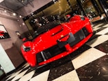 A luxurious red sports car is displayed in a modern showroom with large glass windows and bright lighting. The front of the car features sleek aerodynamic curves, a prominent grille, and distinctive headlights. The flooring consists of a black and white checkered pattern, adding a classic touch to the contemporary setting. Wall art related to cars is visible on the surrounding walls.