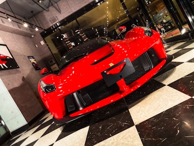 A luxurious red sports car is displayed in a modern showroom with large glass windows and bright lighting. The front of the car features sleek aerodynamic curves, a prominent grille, and distinctive headlights. The flooring consists of a black and white checkered pattern, adding a classic touch to the contemporary setting. Wall art related to cars is visible on the surrounding walls.