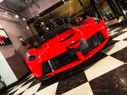 A luxurious red sports car is displayed in a modern showroom with large glass windows and bright lighting. The front of the car features sleek aerodynamic curves, a prominent grille, and distinctive headlights. The flooring consists of a black and white checkered pattern, adding a classic touch to the contemporary setting. Wall art related to cars is visible on the surrounding walls.