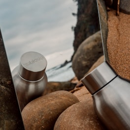 A stainless steel bottle is reflected in a mirror placed among smooth, brown rocks on a sandy beach. The ocean waves and a rocky cliff are visible in the background under a cloudy sky.