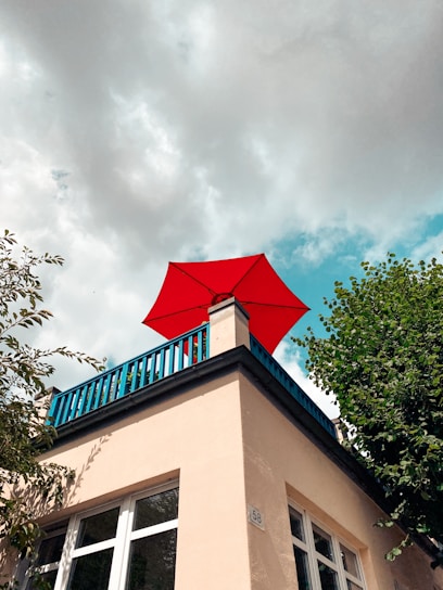 A red umbrella is situated atop a beige building with blue railings. The building features large windows and is surrounded by green foliage. The sky above is partly cloudy, adding a contrasting backdrop to the vibrant colors.