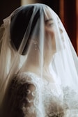 A bride is captured in a soft, intimate moment, partially obscured by a delicate white veil. The intricate lace details of the wedding dress and the elegant earrings add an element of sophistication to the scene.