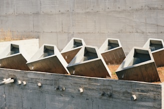 A series of large, angular concrete structures positioned in a uniform row against a backdrop of a textured concrete wall. The structures have a geometric design with sloped edges and hollow interiors, suggesting they may serve an architectural or engineering purpose. Dry grass can be seen around the bases, indicating an outdoor setting.