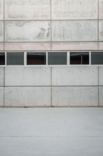 A minimalist and modern architectural facade composed of large, gray concrete panels with visible support bolts. A row of rectangular windows is inserted between two horizontal sections of the wall. The ground is made of smooth concrete, creating a seamless look with the building.