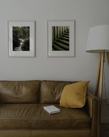 A room with a brown leather sofa, decorated with a yellow pillow. Above the sofa are two framed pictures; one depicts a waterfall in a forest setting, and the other displays a close-up of green leaves. A white floor lamp with a wooden stand is positioned to the side of the sofa. A book with a white cover rests on the sofa.