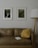 A room with a brown leather sofa, decorated with a yellow pillow. Above the sofa are two framed pictures; one depicts a waterfall in a forest setting, and the other displays a close-up of green leaves. A white floor lamp with a wooden stand is positioned to the side of the sofa. A book with a white cover rests on the sofa.