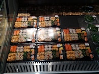 Several packaged trays of assorted sushi are neatly arranged in a refrigerated display. Each tray contains various types of sushi rolls, sashimi, and garnishes, wrapped in plastic for freshness. The trays are aligned in rows, with similar contents across them. Nearby, there are additional packages containing green seaweed salad.