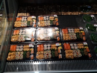 Several packaged trays of assorted sushi are neatly arranged in a refrigerated display. Each tray contains various types of sushi rolls, sashimi, and garnishes, wrapped in plastic for freshness. The trays are aligned in rows, with similar contents across them. Nearby, there are additional packages containing green seaweed salad.