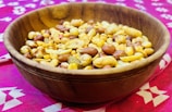A wooden bowl filled with a mixed assortment of nuts and snacks, including peanuts and possibly chickpeas, placed on a vibrant pink patterned fabric.