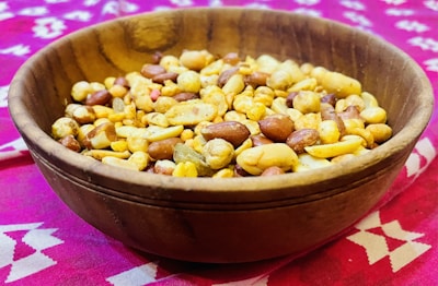 A wooden bowl filled with a mixed assortment of nuts and snacks, including peanuts and possibly chickpeas, placed on a vibrant pink patterned fabric.