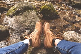 A pair of tan hiking boots worn by someone is resting on rocky terrain beside a flowing stream. The person is wearing rolled-up blue jeans, and the rocks around the feet are covered with patches of moss.
