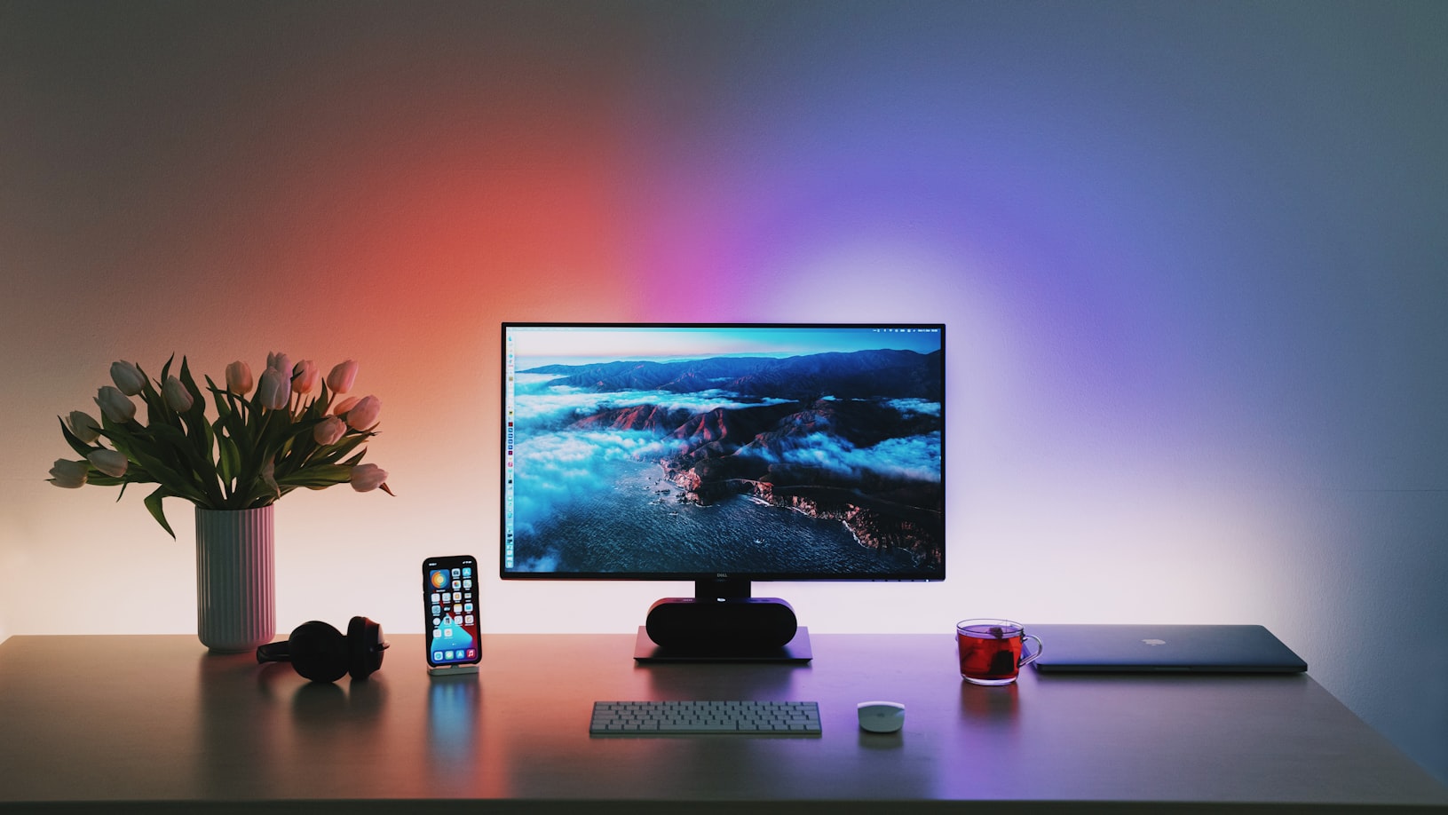 wood table with work stuff on it, such as a monitor displaying a foggy island, a cup of tea, a closed laptop, a white keyboard and mouse, an iphone, a headphone and a jar of flowers, with an overall blue to red gradient light