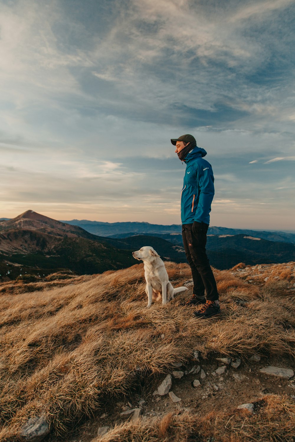 man in blue jacket standing beside white dog on brown grass field during daytime