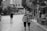 Two people are walking down a cobblestone street, arm in arm, conveying a sense of companionship and intimacy. The scene includes surrounding trees, buildings, and various pedestrians in an urban setting.