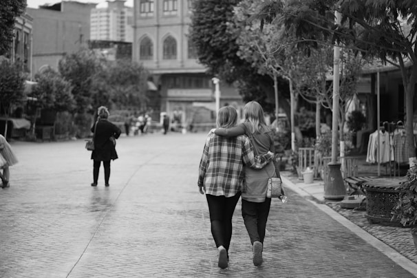Two people are walking down a cobblestone street, arm in arm, conveying a sense of companionship and intimacy. The scene includes surrounding trees, buildings, and various pedestrians in an urban setting.