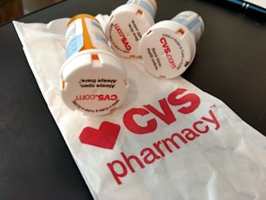 Several prescription pill bottles are lying on a table next to a crumpled paper bag with the CVS Pharmacy logo visible. The tops of the bottles have text for opening instructions and some show the CVS website.