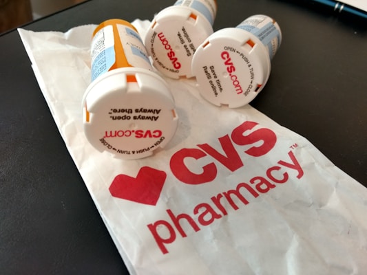 Several prescription pill bottles are lying on a table next to a crumpled paper bag with the CVS Pharmacy logo visible. The tops of the bottles have text for opening instructions and some show the CVS website.