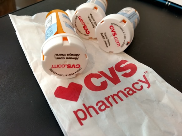Several prescription pill bottles are lying on a table next to a crumpled paper bag with the CVS Pharmacy logo visible. The tops of the bottles have text for opening instructions and some show the CVS website.