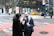 Two people are standing on a city sidewalk near a crosswalk, engaged in conversation. One person, dressed in a dark coat and scarf, is holding a white bag and appears to be speaking. The other person, wearing a dark coat with long hair, is attentive. Behind them, there's a busy street with a yellow taxi and other vehicles passing by. The background features a modern building with large glass windows displaying colorful advertisements.