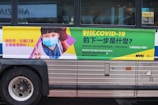 A bus features an advertisement with a message about COVID-19. On the left side, a child wearing a face mask is depicted giving a peace sign. The text, printed in Chinese, discusses COVID-19 prevention and encourages mask-wearing. The right side of the advertisement includes further informational text in green and yellow, with contact details for COVID testing and the NYC and Test & Trace logos.