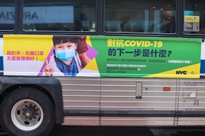 A bus features an advertisement with a message about COVID-19. On the left side, a child wearing a face mask is depicted giving a peace sign. The text, printed in Chinese, discusses COVID-19 prevention and encourages mask-wearing. The right side of the advertisement includes further informational text in green and yellow, with contact details for COVID testing and the NYC and Test & Trace logos.