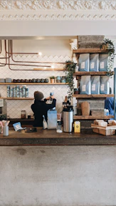 A friendly barista pouring tea behind a small counter decorated with colorful mugs and jars.