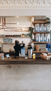A cozy coffee shop counter with a barista preparing a drink. Shelves above the counter hold coffee bags and small plants, creating a warm and inviting atmosphere. The background features a patterned tile wall and copper piping for a modern, industrial design.