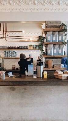 A cozy coffee shop counter with a barista preparing a drink. Shelves above the counter hold coffee bags and small plants, creating a warm and inviting atmosphere. The background features a patterned tile wall and copper piping for a modern, industrial design.