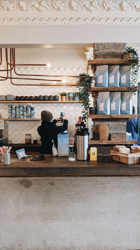 A cozy coffee shop counter with a barista preparing a drink. Shelves above the counter hold coffee bags and small plants, creating a warm and inviting atmosphere. The background features a patterned tile wall and copper piping for a modern, industrial design.