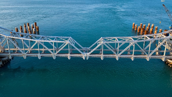 A partially constructed metal bridge extends over a body of calm blue water. The bridge structure is incomplete, with gaps on either end near the water where construction appears to be ongoing. Rusted cylindrical pylons are visible in the water on both sides, possibly for future support or as remnants of previous construction. A crane is seen in the background on the right side, indicating active development.