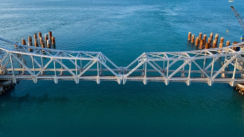 A partially constructed metal bridge extends over a body of calm blue water. The bridge structure is incomplete, with gaps on either end near the water where construction appears to be ongoing. Rusted cylindrical pylons are visible in the water on both sides, possibly for future support or as remnants of previous construction. A crane is seen in the background on the right side, indicating active development.