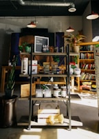 A cozy bookshop corner featuring a display shelf with various items such as coffee beans, snacks, and merchandise. Several potted plants add greenery, while books are neatly arranged on shelves in the background. Warm lighting from hanging lamps overhead gives a comforting ambiance.