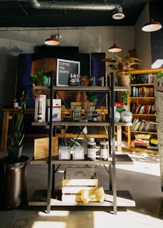 A cozy bookshop corner featuring a display shelf with various items such as coffee beans, snacks, and merchandise. Several potted plants add greenery, while books are neatly arranged on shelves in the background. Warm lighting from hanging lamps overhead gives a comforting ambiance.